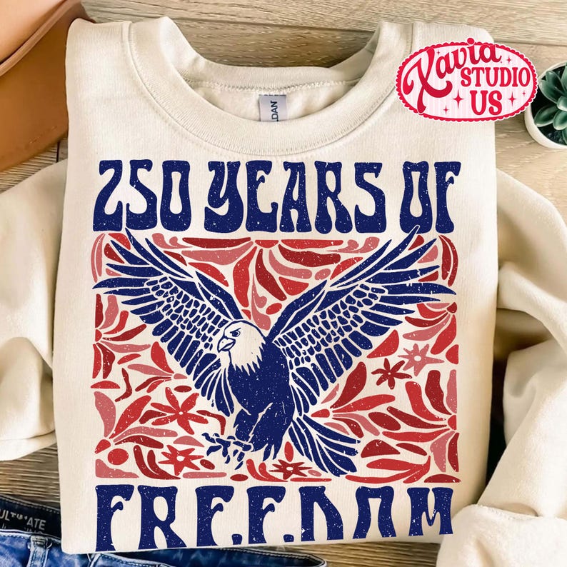 Puede incluir: Sudadera de color crema con un dise&ntilde;o patri&oacute;tico. La parte delantera presenta el texto "250 YEARS OF FREEDOM" en azul, con un &aacute;guila azul y detalles florales rojos. Un logotipo rojo y blanco est&aacute; en la esquina superior derecha.