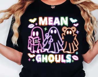 Può includere: T-shirt nera con la scritta "MEAN GHOULS" in colori neon. Il design presenta tre illustrazioni di fantasmi con contorni al neon. Un fantasma legge un libro, un altro tiene una caramella e il terzo ha l'aspetto di un orso.