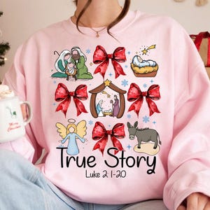 May include: Pink sweatshirt with a Christmas-themed graphic. The design includes illustrations of a nativity scene, red bows, an angel, and a donkey. The text "True Story" and "Luke 2:1-20" are also featured. A festive and religious themed apparel item.