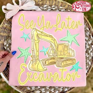 Pu&ograve; includere: T-shirt rosa con grafica di escavatore giallo e la scritta "See Ya Later Excavator". Il design include stelle verdi e blu. Articolo di abbigliamento per bambini.
