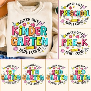May include: A set of six colorful graphic designs for t-shirts featuring the text "Watch Out" and different grade levels, including "Kindergarten", "Preschool", "Pre-K", "1st Grade", "2nd Grade", "3rd Grade", and "4th Grade". Each design includes a pencil, paper airplane, hearts, stars, and flowers.