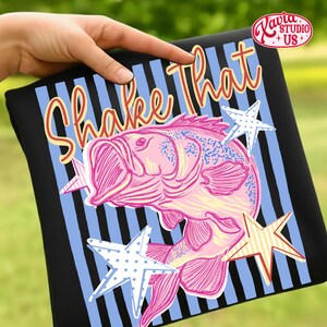 May include: Black t-shirt with a pink fish graphic and the words "Shake That" in a playful font. The design includes blue and white striped background and star accents. The shirt is held by a hand.