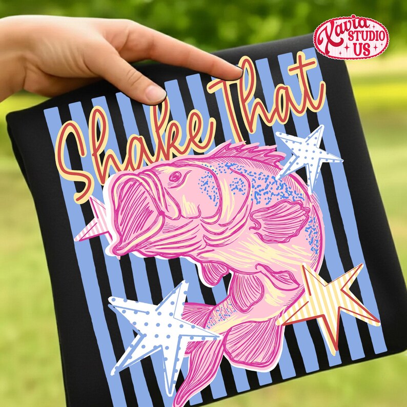 May include: Black t-shirt with a pink fish graphic and the words "Shake That" in a playful font. The design includes blue and white striped background and star accents. The shirt is held by a hand.
