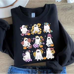 May include: Black sweatshirt featuring a graphic of nine white ghosts adorned with colorful flowers and small pumpkins. The ghosts have black eyes and are surrounded by small star shapes. The sweatshirt is folded on top of blue jeans.