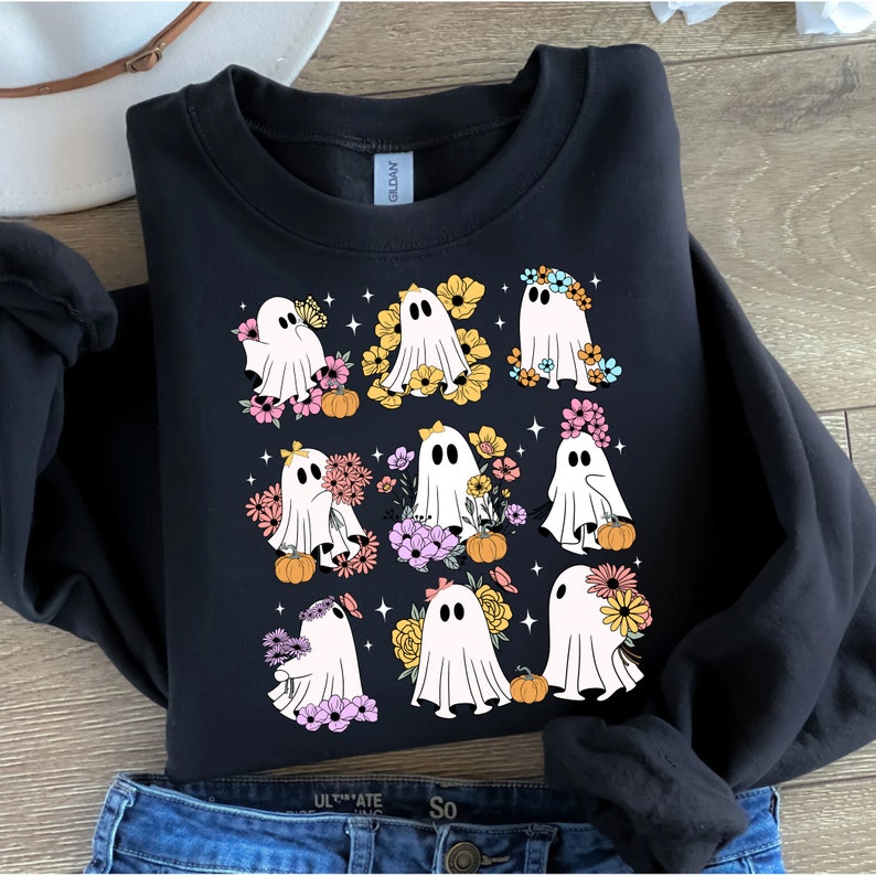 May include: Black sweatshirt featuring a graphic of nine white ghosts adorned with colorful flowers and small pumpkins. The ghosts have black eyes and are surrounded by small star shapes. The sweatshirt is folded on top of blue jeans.