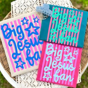 May include: Three colorful tote bags with the text "Big Jesus Fan" in a playful font. The bags are in beige, teal, and pink, with vertical stripes and star accents. A light blue hair clip and a necklace are on top.