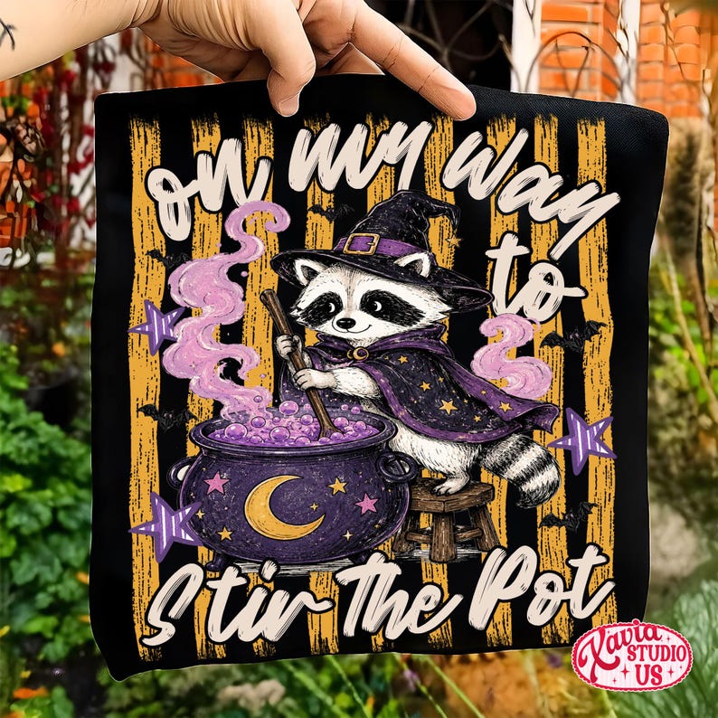 Peut inclure: Un article en tissu noir avec un motif de raton laveur portant un chapeau de sorci&egrave;re et une cape, remuant un chaudron violet. Le texte indique "on my way to STIR THE POT" sur fond de rayures orange, d'&eacute;toiles violettes et de chauves-souris.