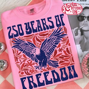 Puede incluir: Camiseta rosa con un dise&ntilde;o gr&aacute;fico. Presenta un &aacute;guila azul con las alas extendidas, rodeada de elementos florales rojos y rosas. El texto "250 YEARS OF FREEDOM" est&aacute; impreso en azul, con un estilo vintage.