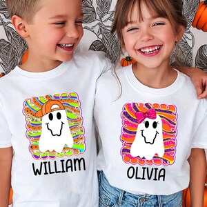 May include: Two children wearing white t-shirts with Halloween ghost designs. One ghost wears an orange cap, the other a pink bow. The shirts have the names "WILLIAM" and "OLIVIA" printed below the ghosts. The background is a leaf pattern.