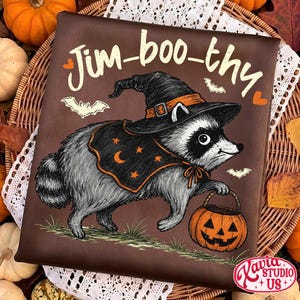 May include: A dark brown t-shirt featuring a graphic of a raccoon wearing a black witch hat and a cape with orange stars and a crescent moon. The raccoon holds an orange jack-o'-lantern bucket. The text Jim-boo-thy is printed above.