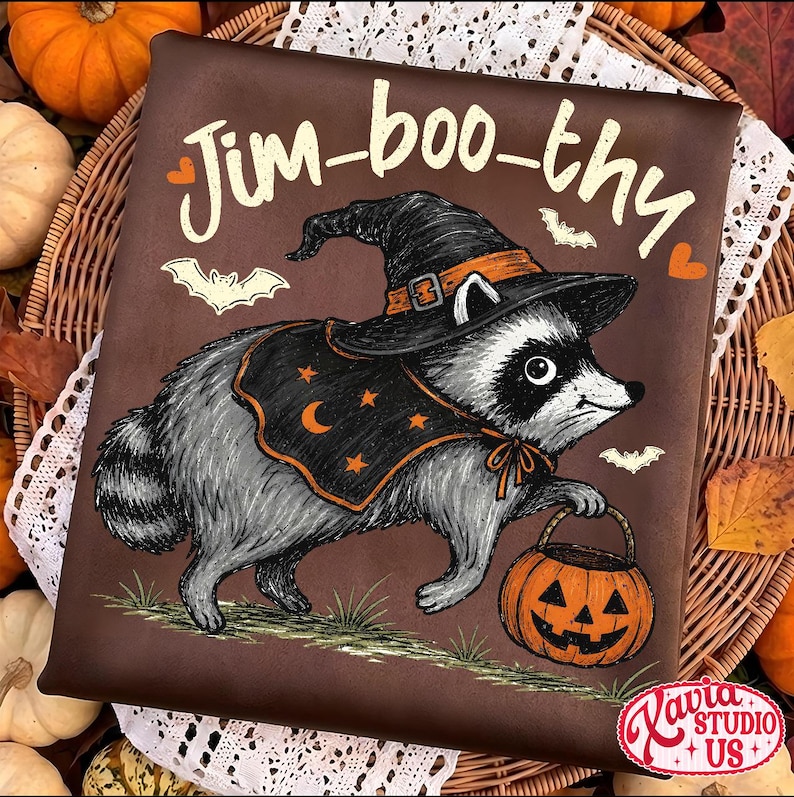 May include: A dark brown t-shirt featuring a graphic of a raccoon wearing a black witch hat and a cape with orange stars and a crescent moon. The raccoon holds an orange jack-o'-lantern bucket. The text Jim-boo-thy is printed above.