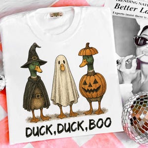 May include: White t-shirt featuring three ducks dressed in Halloween costumes. One duck is a witch, one is a ghost, and one is a jack-o'-lantern. The text "DUCK, DUCK, BOO" is printed below the ducks.