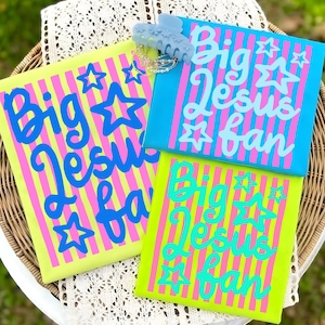 May include: Three colorful canvas pouches with the text "Big Jesus Fan" in a playful font. The pouches are in bright colors: yellow, blue, and green, with pink and blue vertical stripes. A light blue hair clip and a bracelet are on top.