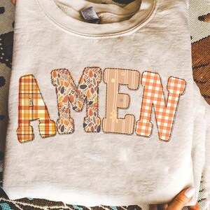 Puede incluir: Una sudadera color crema con la palabra "AMEN" en letras grandes y decorativas. Las letras están en varios patrones temáticos de otoño, incluyendo cuadros, florales y vichy, en tonos naranja, marrón y beige.