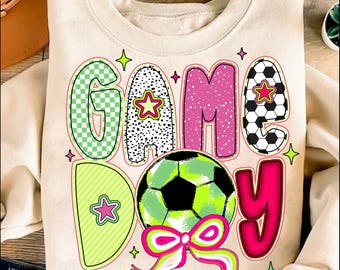 Puede incluir: Una sudadera de color crema con un diseño gráfico colorido que dice "GAME DAY" con un balón de fútbol en el medio. Las letras están hechas de diferentes patrones y colores, incluyendo rosa, verde y negro.