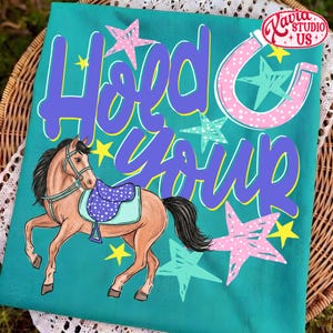 May include: Teal t-shirt with a horse illustration and the text "Hold Your Horses" in purple and white. The horse has a saddle and is surrounded by stars in pink, teal, and yellow. A horseshoe graphic is also present.