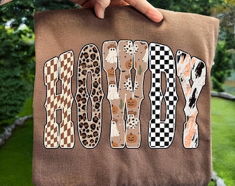 May include: A brown sweatshirt featuring the word "HOWDY" in a decorative font. The letters are filled with patterns such as a checkerboard, leopard print, and Halloween-themed designs including ghosts and pumpkins.