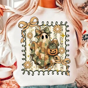 May include: White sweatshirt featuring a Halloween ghost design. The ghost is wearing a camouflage sheet with a bow and holding a pumpkin basket. The design includes flowers, bows, and a decorative border.