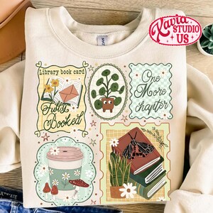 May include: Cream-colored sweatshirt with a colorful graphic design featuring book-themed illustrations. The design includes a coffee cup, books, plants, and the phrases "Fully Booked" and "One More Chapter."