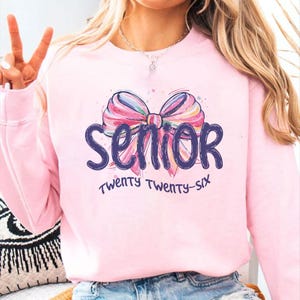 May include: A light pink sweatshirt with a colorful bow graphic and the word "Senior" in dark blue script. Below the word "Senior" is the text "Twenty Twenty-Six" in a handwritten style font.