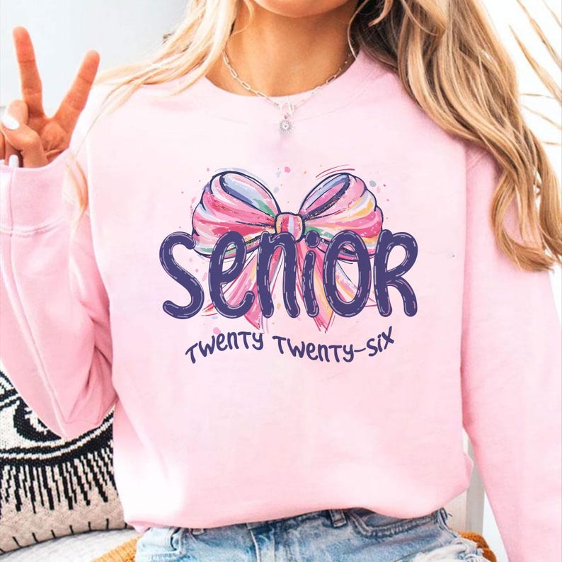 May include: A light pink sweatshirt with a colorful bow graphic and the word "Senior" in dark blue script. Below the word "Senior" is the text "Twenty Twenty-Six" in a handwritten style font.