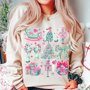 May include: Beige sweatshirt with a festive Christmas design. The design includes a sleigh, bows, a Christmas tree, presents, a nutcracker, and a floral ornament. The illustrations are in shades of pink, green, and white.