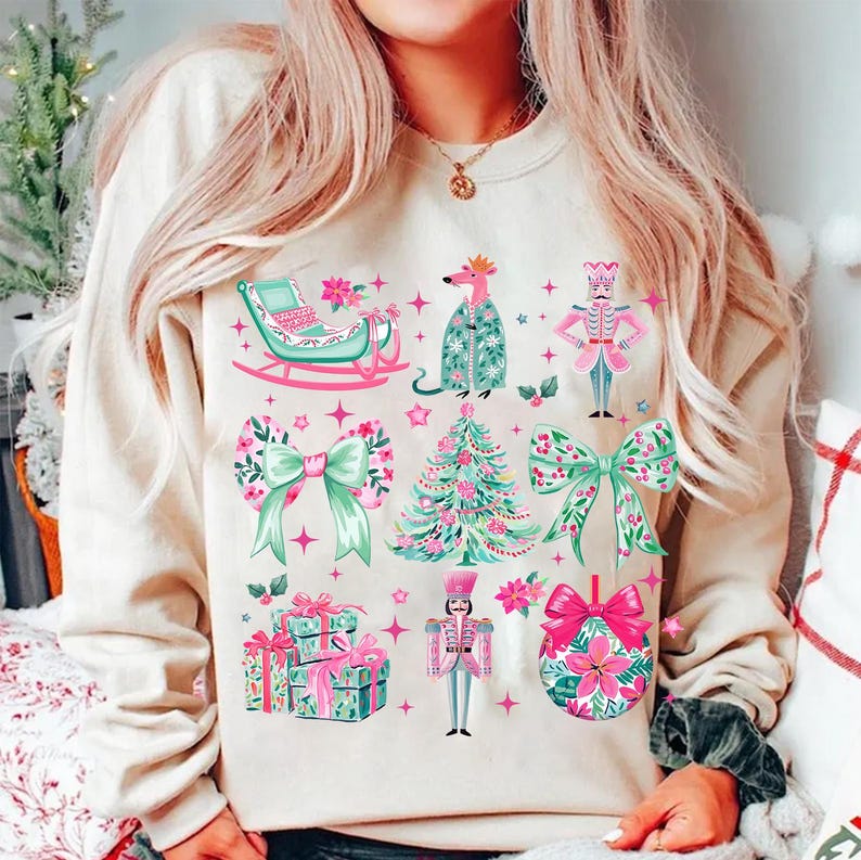 May include: Beige sweatshirt with a festive Christmas design. The design includes a sleigh, bows, a Christmas tree, presents, a nutcracker, and a floral ornament. The illustrations are in shades of pink, green, and white.