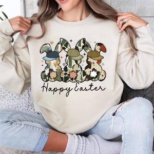 May include: A beige sweatshirt with a graphic of three cartoon bunnies wearing hats and the text "Happy Easter".