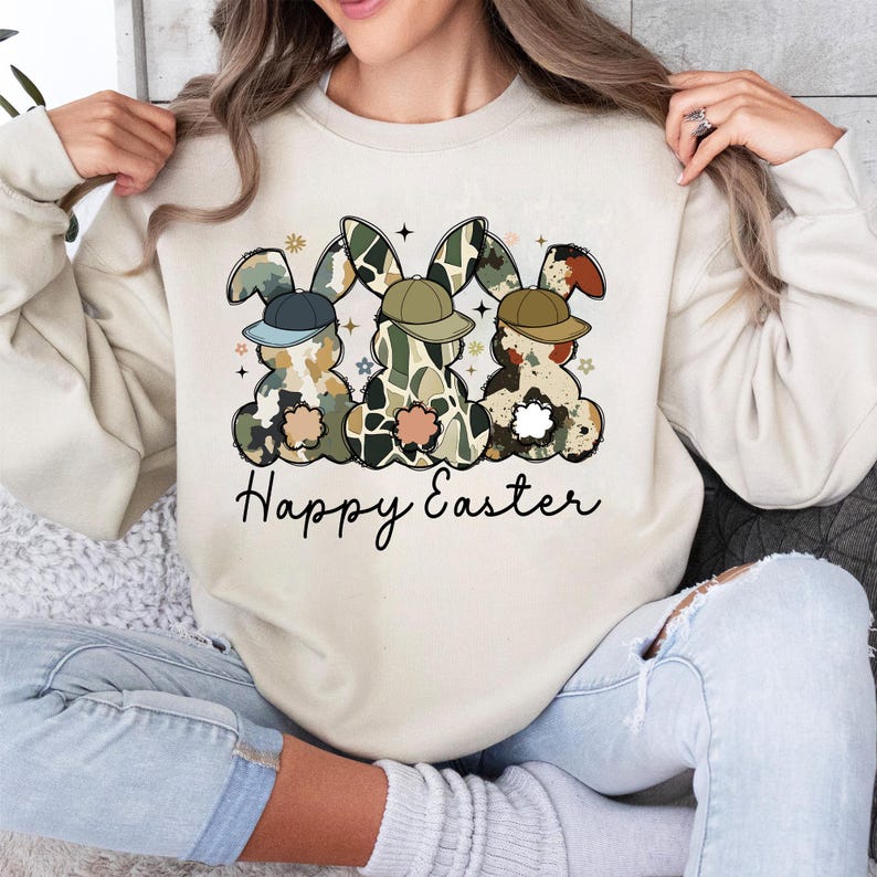 May include: A beige sweatshirt with a graphic of three cartoon bunnies wearing hats and the text "Happy Easter".
