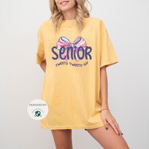 May include: Mustard yellow t-shirt with a colorful bow graphic and the word "Senior" in blue script. Below the word "Senior" is the text "Twenty Twenty-Six" in blue. The shirt is a relaxed fit.