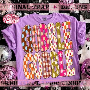 May include: A light purple t-shirt featuring the text GOBBLE GOBBLE in large, multi-patterned letters. The letters include designs such as blue polka dots, red plaid, pink polka dots, orange and white checkers, and white stars on a gold background.