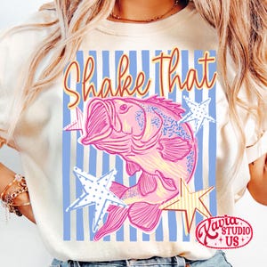May include: Cream-colored t-shirt featuring a pink and blue illustration of a fish with the text "Shake That." The design includes blue and white striped background and star accents. The shirt has a round neckline and short sleeves.
