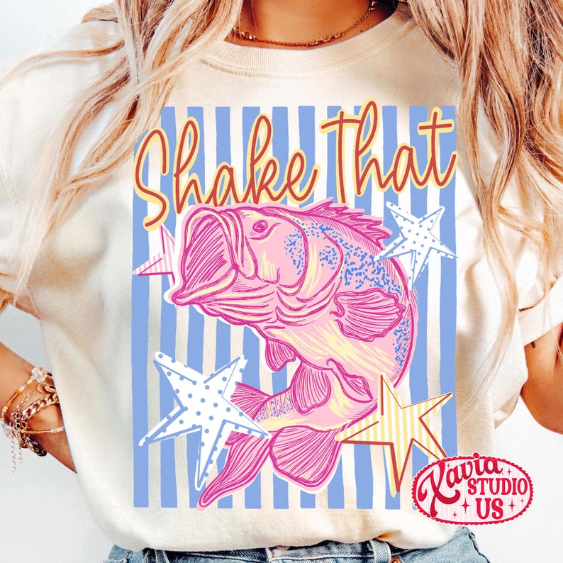 May include: Cream-colored t-shirt featuring a pink and blue illustration of a fish with the text "Shake That." The design includes blue and white striped background and star accents. The shirt has a round neckline and short sleeves.