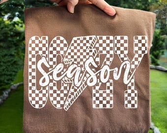May include: A brown tote bag with the words "GOATY Season" in white. The letters "GOATY" are designed with a checkered pattern, and the word "Season" is in a cursive font. The bag is held up by a hand.