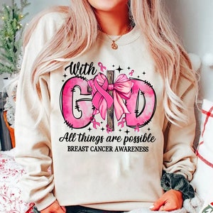 May include: A beige sweatshirt with a pink graphic supporting breast cancer awareness. The graphic includes the words "With God, All things are possible" and a pink ribbon. The sweatshirt has a crew neck.