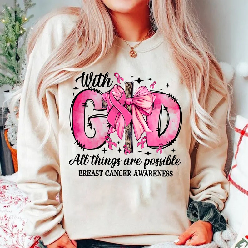 May include: A beige sweatshirt with a pink graphic supporting breast cancer awareness. The graphic includes the words "With God, All things are possible" and a pink ribbon. The sweatshirt has a crew neck.