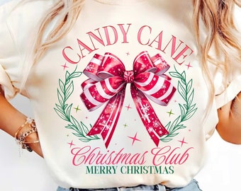 May include: Cream-colored sweatshirt featuring a festive design. The graphic includes a large red and white striped bow, surrounded by a green wreath and the words "CANDY CANE CHRISTMAS CLUB MERRY CHRISTMAS" in pink script.
