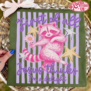 May include: Green t-shirt with a pink raccoon illustration holding a flower, surrounded by stars and vertical stripes. Text reads "First of all Jesus thinks I'm a delight."