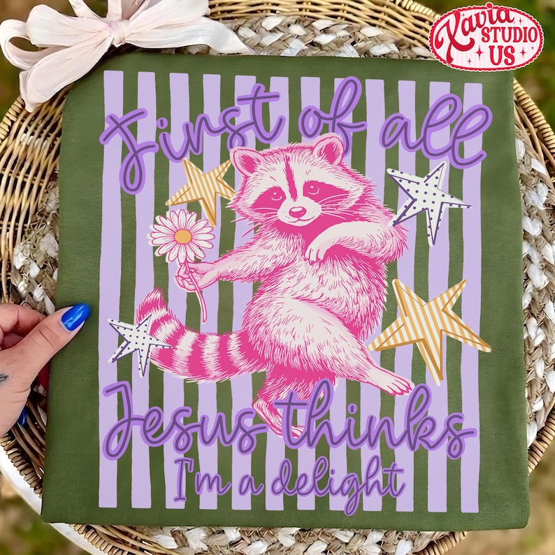 May include: Green t-shirt with a pink raccoon illustration holding a flower, surrounded by stars and vertical stripes. Text reads "First of all Jesus thinks I'm a delight."
