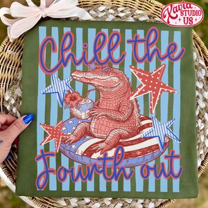May include: Olive green t-shirt with a Fourth of July theme. The design features an alligator wearing sunglasses and floating on an inner tube, holding a tropical drink. The text "Chill the Fourth out" is written in red and blue.