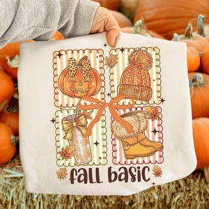 May include: A cream-coloured graphic tee featuring four autumn-themed illustrations: a jack-o'-lantern, a knitted hat, a beverage, and boots. The words "fall basic" are printed at the bottom, with autumn leaves. The illustrations are in orange, brown, and yellow.