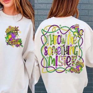 May include: Cream-coloured sweatshirt with a colourful Mardi Gras design. The front features a crown and fleur-de-lis graphic. The back displays the phrase "Throw Me Something Mister" in green, yellow, and purple, surrounded by beaded necklaces.