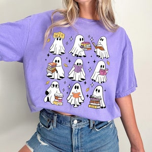 May include: A light purple t-shirt featuring a grid of cartoon ghosts, each holding or reading books. The ghosts are white with black eyes and are surrounded by stars and autumn leaves. The shirt is a comfortable, casual style.