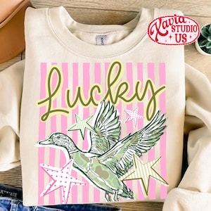 May include: Cream-colored sweatshirt with a graphic design featuring the word "Lucky" in a script font, a duck illustration, and star accents. The background has pink vertical stripes. The sweatshirt has a crew neck.