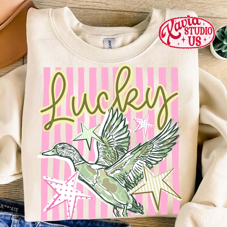 May include: Cream-colored sweatshirt with a graphic design featuring the word "Lucky" in a script font, a duck illustration, and star accents. The background has pink vertical stripes. The sweatshirt has a crew neck.