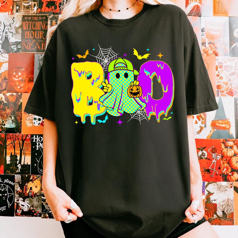 Puede incluir: Camiseta negra con un gr&aacute;fico colorido de "BOO". La "B" es amarilla, la "O" es un fantasma con una gorra verde y la otra "O" es morada. El fantasma sostiene una calabaza. Tambi&eacute;n se presentan murci&eacute;lagos y telara&ntilde;as.