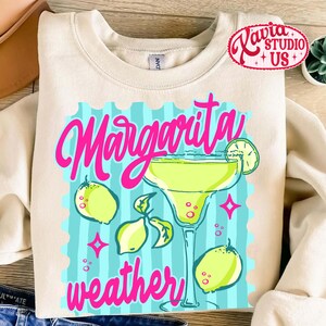 May include: Cream-coloured sweatshirt with a margarita-themed graphic. The design features a margarita glass, lemons, and the words "Margarita weather" in pink. The background is light blue with vertical stripes.