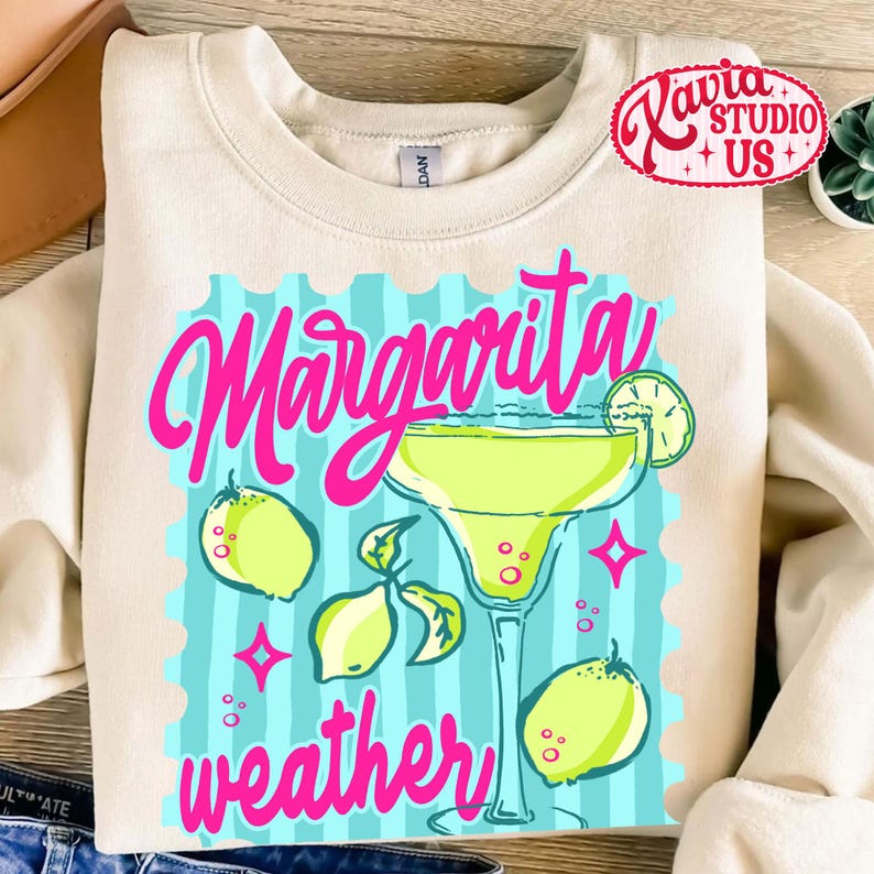 May include: Cream-coloured sweatshirt with a margarita-themed graphic. The design features a margarita glass, lemons, and the words "Margarita weather" in pink. The background is light blue with vertical stripes.
