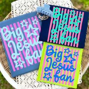 May include: Three colorful graphic t-shirts with the text "Big Jesus Fan" in a script font, accented with stars. The shirts are in navy, blue, and neon green, with pink and blue stripes. A light blue hair clip and jewelry are also in the image.