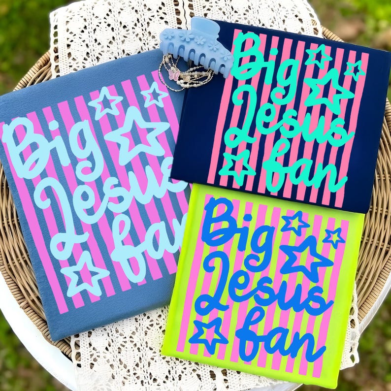 May include: Three colorful graphic t-shirts with the text "Big Jesus Fan" in a script font, accented with stars. The shirts are in navy, blue, and neon green, with pink and blue stripes. A light blue hair clip and jewelry are also in the image.